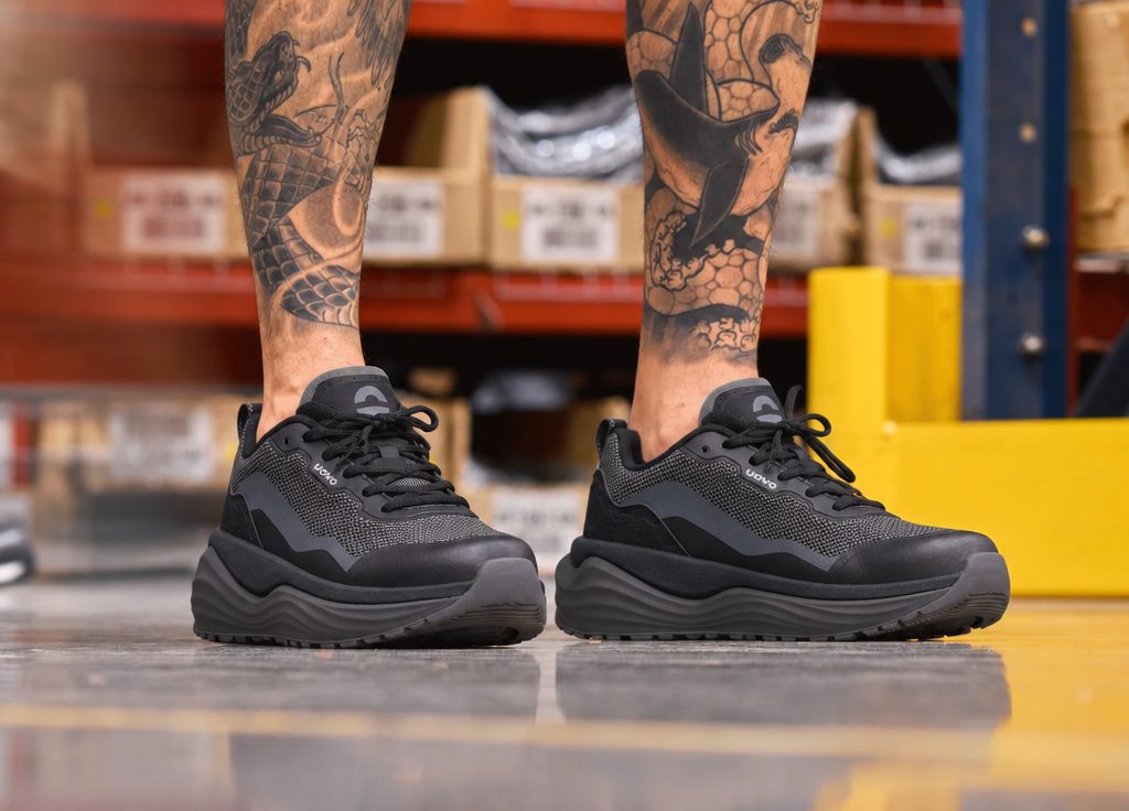 Close-up of black UOVO Flow sneakers on a polished warehouse floor in front of stocked shelves