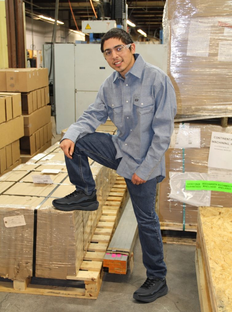 Model in a work shirt and black sneakers posing beside stacked pallets in a warehouse